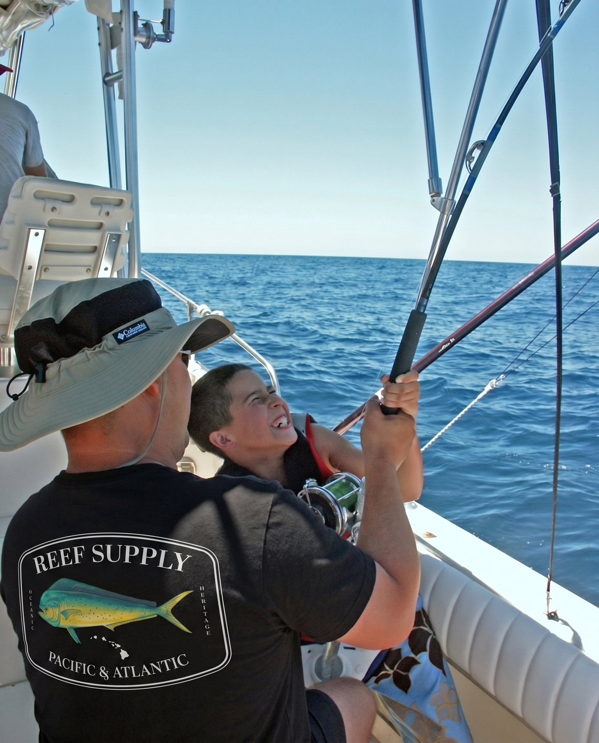 Man in a Reef Supply t-shirt and a child fishing from a boat on the open ocean.
